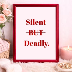 Puede incluir: Una impresión enmarcada con las palabras "Silent BUT Deadly" en letras rojas. El marco rojo rodea un fondo blanco. La imagen está sobre una superficie blanca y esponjosa con una vela y flores.