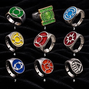 Könnte beinhalten: Eine Sammlung von neun silberfarbenen Metallringen, die jeweils ein einzigartiges farbiges Emaille-Emblem mit verschiedenen Symbolen tragen. Die Ringe sind von eins bis neun nummeriert und zeigen diverse kreisförmige und rechteckige Designs in Grün, Blau, Gelb, Rot und Orange.