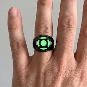 May include: A black metal ring featuring a circular emblem with a glowing neon green centre. The emblem displays a stylised black lantern symbol within the luminous green circle, creating a high-contrast design on the dark band surface.