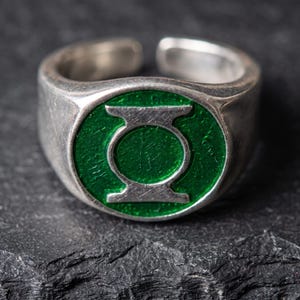 May include: A silver-toned metal ring featuring an open band design. The circular face of the ring displays a vibrant green enamel inlay with a raised silver-colored symbol resembling a stylized lantern logo centered within the green circle.