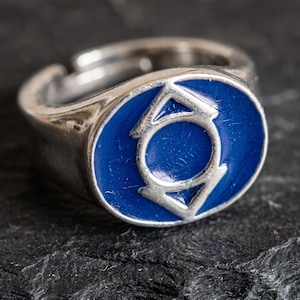 May include: A silver-toned metal ring featuring a circular face with a deep blue enamel inlay. The centre of the blue circle displays a raised silver-coloured symbol consisting of a central circle enclosed by two opposing arrowheads pointing outward.