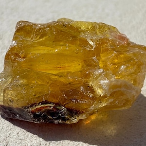 Puede incluir: Un espécimen mineral en bruto, translúcido y de color ámbar con una estructura cristalina irregular. La superficie presenta facetas desiguales, fracturas internas y tonos variables de amarillo dorado, con una inclusión oscura y opaca en la base.