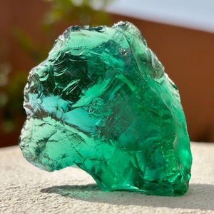 May include: A translucent, emerald green glass rock with a jagged, irregular shape. The surface features sharp, conchoidal fractures and smooth, curved edges that catch the light, creating deep green tones and bright highlights throughout the piece.
