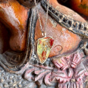 Puede incluir: Un collar de plata con un colgante de piedra translúcida de forma irregular. La piedra tiene inclusiones amarillas y rojas, engastada en un bisel de plata. La cadena es fina, y el colgante cuelga sobre un fondo ornamentado borroso.
