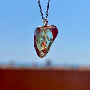 Puede incluir: Un collar con colgante que presenta un dije de resina transparente de forma irregular en corazón, engastado en un marco de metal plateado. El dije contiene fragmentos rojos y amarillos incrustados. El colgante cuelga de una cadena de metal oscuro.