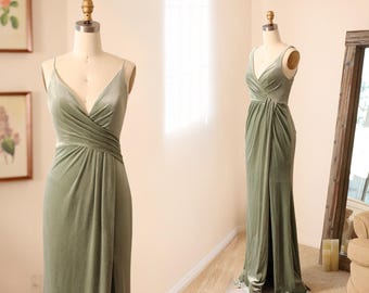 May include: Sage green velvet gown with a V-neckline and spaghetti straps. The dress features a draped bodice and a flowing skirt. The gown is displayed on a mannequin, showcasing its elegant design and silhouette. The dress is floor-length.