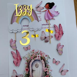 May include: A cake topper featuring the name "Izzy" in gold foil, a rainbow, clouds, and the number "3". It includes a fairy, butterflies, and a fairy door with floral details. Designed for a birthday cake.