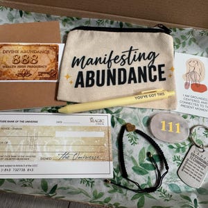 May include: A gift box containing a beige canvas pouch with the text 'manifesting ABUNDANCE', a yellow pen labelled 'YOU'VE GOT THIS', a 'Divine Abundance 888' card, a 'Gratitude Bank of the Universe' cheque, a '111' stone, a black cord bracelet, and a grounding keychain.