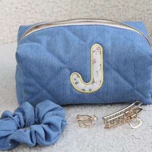 Op de afbeelding: Een blauwe denim quilted toilettas met een gouden rits en een geborduurde bloemletter "J" aan de voorkant. De tas ligt naast een blauwe denim scrunchie en twee gouden haarclips.
