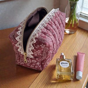 May include: A quilted, burgundy cosmetic bag with a floral pattern and white lace trim. The bag has a gold zipper and is open, revealing a dark interior. A bottle of perfume and a tube of lip balm sit nearby.