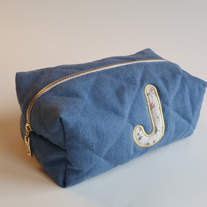 Op de afbeelding: Een blauwe, gewatteerde toilettas met een bloemige stoffen letter "J" aan de voorkant.