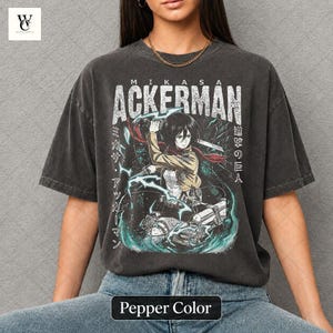 Könnte beinhalten: Pfefferfarbenes T-Shirt mit einer Grafik von Mikasa Ackerman aus Attack on Titan. Das Design beinhaltet den Namen "ACKERMAN" in großen weißen Buchstaben, japanischen Text und eine Illustration der Figur in Aktion. Das Shirt hat einen Vintage-Stil.