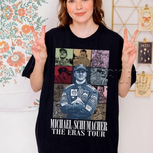 Peut inclure: T-shirt noir avec un collage d'images de Michael Schumacher et le texte "MICHAEL SCHUMACHER THE ERAS TOUR" en blanc. Le t-shirt a un col rond et des manches courtes.
