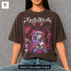 Może przedstawiać: Ciemnobrązowy t-shirt z grafiką z anime Black Clover. Projekt zawiera japoński tekst i żywą ilustrację postaci z mieczem i fioletową energią. Koszulka ma wygląd vintage.