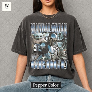 May include: Pepper color t-shirt featuring a graphic of characters from The Mandalorian series. The design includes the text "MANDALORIAN" and "GROGU" in a metallic blue font, along with images of Grogu and Mandalorian characters.