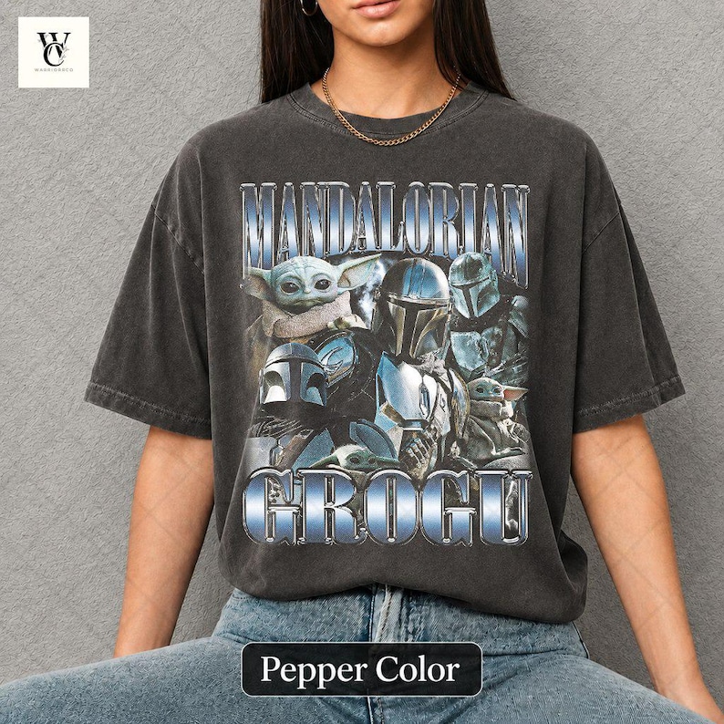 May include: Pepper color t-shirt featuring a graphic of characters from The Mandalorian series. The design includes the text "MANDALORIAN" and "GROGU" in a metallic blue font, along with images of Grogu and Mandalorian characters.