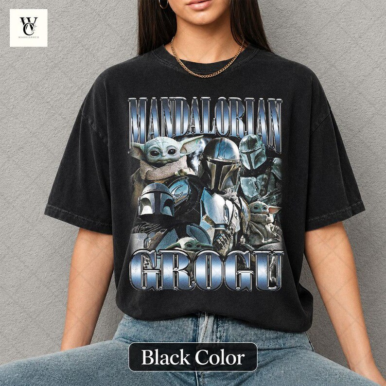 May include: Black t-shirt featuring the text "MANDALORIAN GROGU" in a metallic blue font. The graphic includes images of characters from the Star Wars series. The shirt is a dark color and has a relaxed fit.