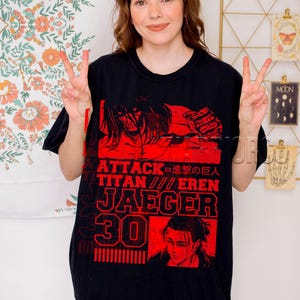 Op de afbeelding: Zwart T-shirt met een rode grafische print van personages uit de anime Attack on Titan. De tekst op het shirt luidt "Attack on Titan // Eren Jaeger 30".