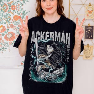Pu&ograve; includere: T-shirt nera con grafica che presenta il nome "Levi Ackerman" in lettere bianche invecchiate. Il design include un'illustrazione di un personaggio con una spada, testo giapponese e uno sfondo turchese e bianco.