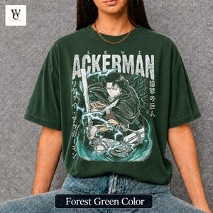 Pu&ograve; includere: T-shirt verde foresta con grafica di un personaggio con una spada, il nome "ACKERMAN" in bianco e testo giapponese. Il design include effetti di fulmini e acqua. La maglietta ha una vestibilit&agrave; ampia.
