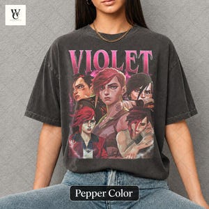 以下が含まれることがあります： ピンク色の「VIOLET」の文字と、複数のキャラクターのグラフィックが特徴の濃いグレーのTシャツ。 シャツはペッパーカラーで、ヴィンテージ感のある着古した外観です。 デザインには、赤と黒の髪の複数の人物が含まれています。