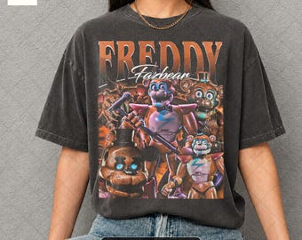 May include: Pepper color t-shirt featuring a graphic of Freddy Fazbear characters. The design includes the text "Freddy Fazbear" in orange, with various characters in the center. The shirt is a dark gray color.