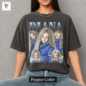 May include: Pepper color t-shirt featuring a graphic of a woman with blonde hair in a blue outfit, with the name "DIANA" in blue text above. The shirt has a vintage, distressed look.
