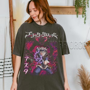 Może przedstawiać: Ciemnoszara koszulka z grafiką postaci z anime z fioletowymi efektami energetycznymi. Koszulka ma japoński tekst na g&oacute;rze i imię postaci po japońsku po lewej stronie. Koszulka jest noszona przez osobę.