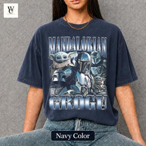 May include: Navy blue t-shirt featuring a graphic of characters from The Mandalorian series, including Grogu and Mandalorian figures. The text "MANDALORIAN GROGU" is displayed in a metallic, blue font. The shirt is a relaxed fit.