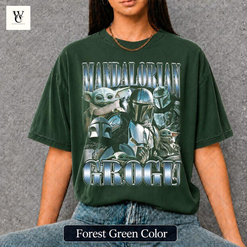 May include: Forest green t-shirt featuring a graphic of characters from The Mandalorian, including Grogu and the Mandalorian. The text "MANDALORIAN" and "GROGU" are displayed in a metallic, blue-silver font. The shirt is a relaxed fit.