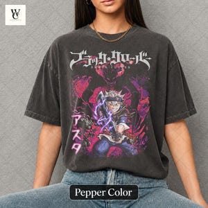 Może przedstawiać: Koszulka w kolorze pieprzu z grafiką z anime Black Clover. Projekt zawiera japoński tekst i ilustrację postaci z fioletowymi efektami błyskawic. Koszulka jest w kolorze ciemnoszarym.