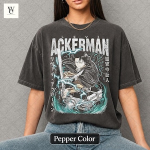 Pu&ograve; includere: T-shirt color pepe con grafica di un personaggio con una spada, il nome "ACKERMAN" in bianco e testo giapponese. Il design include accenti turchese e bianchi. La maglietta &egrave; in stile vintage.