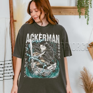 Pu&ograve; includere: T-shirt grigio scuro con la scritta "ACKERMAN" sopra un'illustrazione in stile anime di un personaggio con una spada. La grafica include testo giapponese e dettagli turchese. La maglietta &egrave; indossata da una persona.