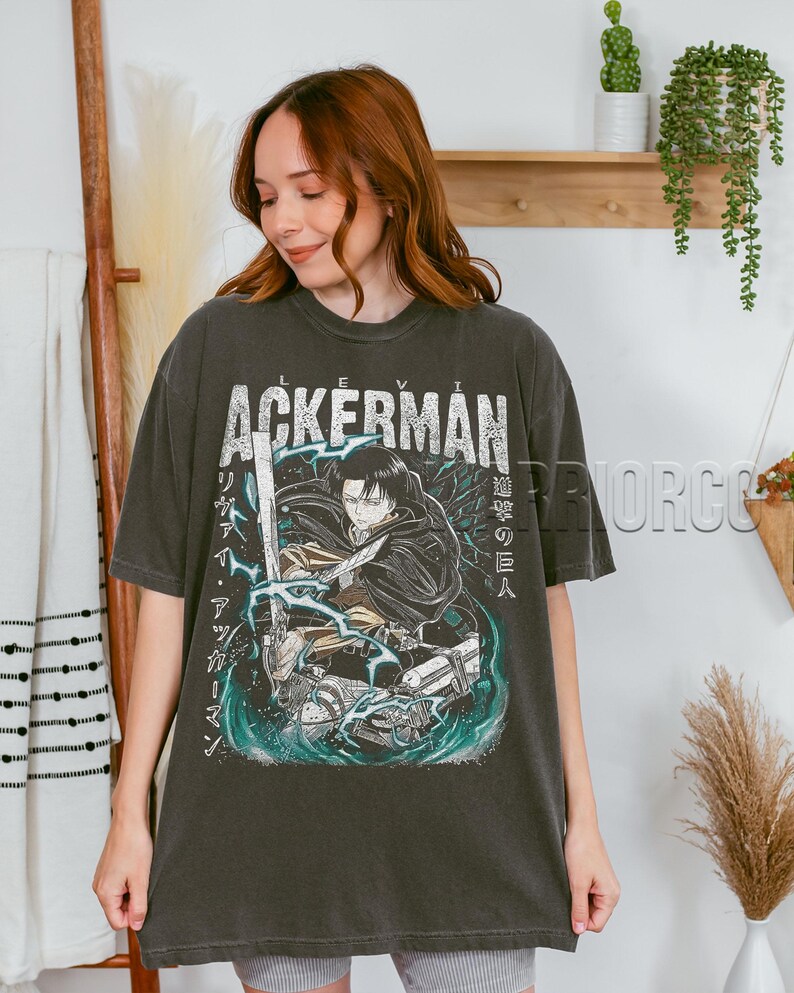 Pu&ograve; includere: T-shirt grigio scuro con la scritta "ACKERMAN" sopra un'illustrazione in stile anime di un personaggio con una spada. La grafica include testo giapponese e dettagli turchese. La maglietta &egrave; indossata da una persona.