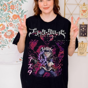 Puede incluir: Camiseta negra con un diseño gráfico de la serie de anime Black Clover. El diseño incluye el título de la serie en japonés e inglés, junto con una ilustración de personaje en morado y blanco.