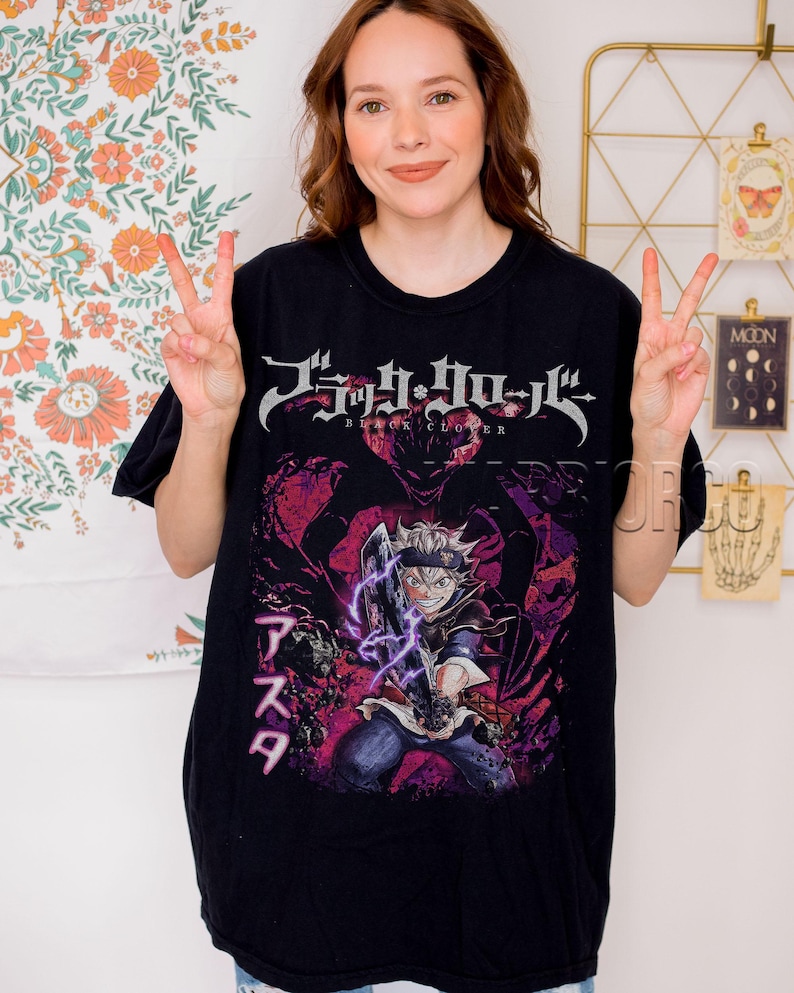 Może przedstawiać: Czarny t-shirt z grafiką z anime Black Clover. Projekt zawiera tytuł serii w kolorze srebrnym i japoński tekst, a także ilustrację postaci w odcieniach fioletu i czerwieni.