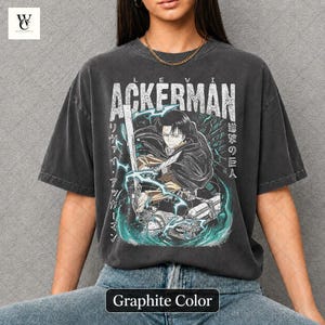 Pu&ograve; includere: T-shirt color grafite con grafica di un personaggio con una spada, testo giapponese e il nome "ACKERMAN". Il design include dettagli turchese e bianchi. La maglietta ha un aspetto vintage e usurato.