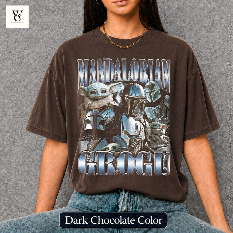 May include: Dark chocolate-colored t-shirt featuring the text "MANDALORIAN GROGU" in a metallic blue font. The graphic includes images of characters from the Star Wars series, including Grogu and Mandalorian armor. The shirt is a relaxed fit.