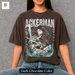Pu&ograve; includere: T-shirt color cioccolato fondente con grafica di un personaggio con una spada, il nome "ACKERMAN" in bianco e testo giapponese. Il design include fulmini turchesi e un'illustrazione dettagliata.