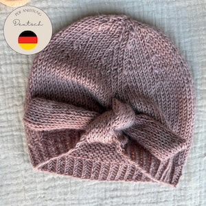 Könnte beinhalten: Ein gestrickter mauvefarbener Baby-Turban mit geknoteter Vorderseite. Der Hut ist aus weichem, strukturiertem Garn gefertigt und hat einen gerippten Saum. Das Bild enthält Text auf Deutsch.