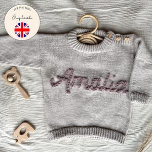 Peut inclure: Un pull pour bébé en tricot gris clair avec le nom "Amalia" brodé en mauve. Le pull est sur un cintre en bois, avec deux jouets en bois et un logo "PDF Pattern English". L'image est sur une surface texturée gris clair.