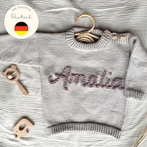 May include: A light gray knitted baby sweater with the name "Amalia" embroidered in a mauve color. The sweater has wooden buttons on the shoulder and is displayed on a wooden hanger. Wooden baby toys are also visible.