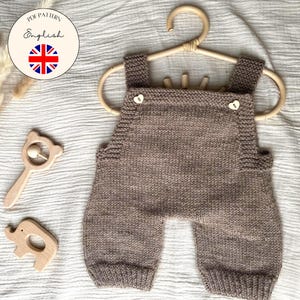 Könnte beinhalten: Eine gestrickte braune Baby-Latzhose mit herzförmigen Knöpfen, die an einem Holzbügel hängt. Daneben befinden sich zwei Holzspielzeuge: eine bärenförmige Rassel und ein Elefant. Der Text "PDF Pattern English" und "Children & Baby Knitting Pattern" ist ebenfalls sichtbar.
