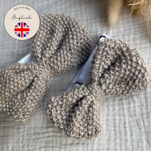 Könnte beinhalten: Zwei beige gestrickte Haarschleifen. Die Schleifen bestehen aus einem grob gestrickten Material und sind an weißen Clips befestigt. Das Bild enthält eine runde Grafik mit dem Text "PDF Pattern English" und einer britischen Flagge. Der Text "MASCHENMINIS - KINDER & BABYSTRICK ANLEITUNGEN" ist ebenfalls sichtbar.