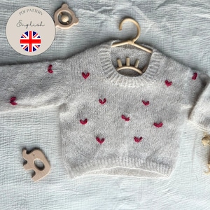 Könnte beinhalten: Ein hellgrauer Baby-Pullover mit roten herzförmigen Stickereien. Der Pullover wird auf einem Holzkleiderbügel präsentiert. Das Bild enthält auch Holzspielzeug und den Text "PDF Pattern English".