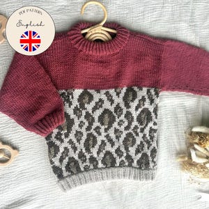 May include: A knitted baby sweater in shades of burgundy, gray, and white, featuring a leopard print design on the lower half. The sweater is displayed on a wooden hanger. A wooden toy and a round sticker with the Union Jack flag are also visible.