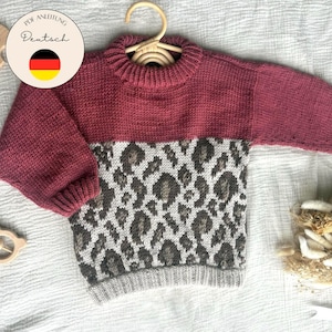 May include: A knitted baby sweater in shades of burgundy, gray, and white, with a leopard print design on the front. The sweater is displayed on a wooden hanger. The image also includes a wooden rattle and a sign that says "PDF Anleitung Deutsch".