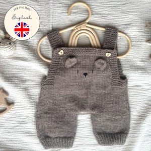 Könnte beinhalten: Graue gestrickte Baby-Latzhose mit Bärenohren und einem schwarzen, gestickten Gesicht. Die Latzhose hängt an einem Holzkleiderbügel. Ein Holzspielzeug und ein gestrickter Hase sind ebenfalls im Bild. Der Text "English" und eine UK-Flagge sind in einem Kreis.