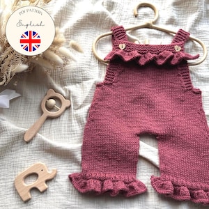 Puede incluir: Un pelele de bebé tejido, de color rosa polvoriento, con bordes con volantes y botones de madera, cuelga de una percha de madera. Un sonajero y un juguete de elefante de madera están cerca. El texto dice "PDF Pattern English".