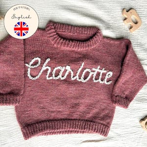 Könnte beinhalten: Ein altrosa Strickpullover mit dem Namen "Charlotte" in Weiß gestickt. Der Pullover hat einen gerippten Kragen und Bündchen. Das Bild enthält auch Holzspielzeug und den Text "Children & Baby Knitting Pattern".